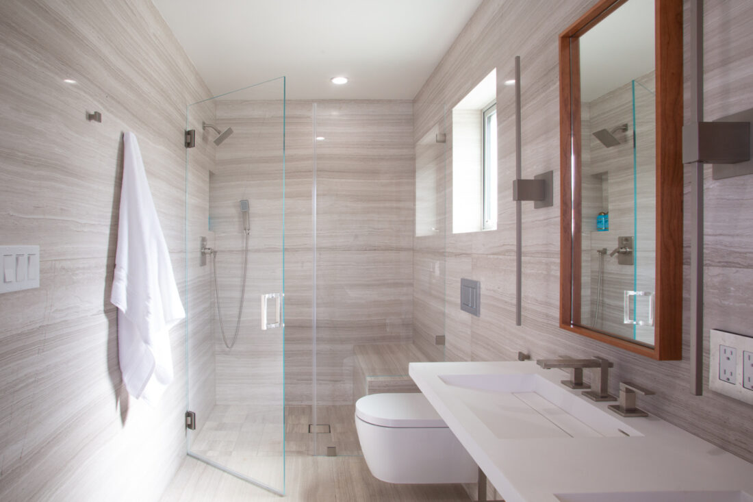 A modern bathroom remodel with sandstone slab walls and a glass shower and sink.